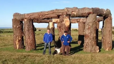 A picture of Nick Gray and Charlie Newman with a dog between them in front of the woodhenge creation.