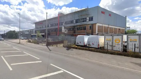 Queen Anne Works Holyhead Road. It is a large factory with scaffolding on the outside. It is a busy main road. There are vans and lorries on a nearby parking lot. 