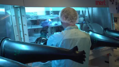 A scientist dressed in a blue lab coat with their arms through black gloves into a glass container