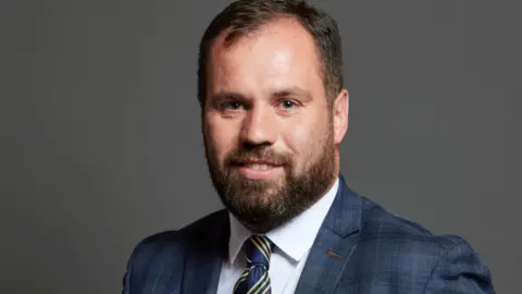 UK Parliament Sean Woodcock's parliamentary photo portrait. He has short brown hair and a brown beard. He wears a blue suit, blue and gold striped tie, and white shirt.