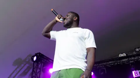 Bemz performs onstage, while wearing a white shirt and green shorts. He is holding a microphone in one hand.