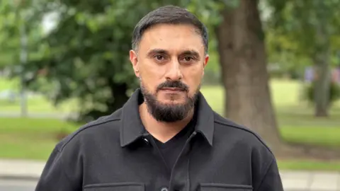 Amjid Khazir is stood in a black jacket near a park in Middlesbrough