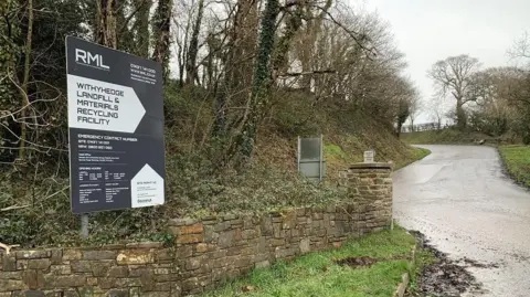 Sign for Withyhedge landfill on the left by a wall and trees with a path/road to the right