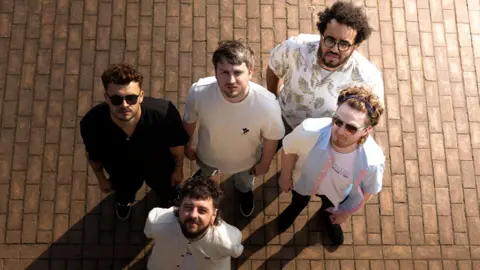 Leazes The five members of Leazes are looking up at the camera above them. Four of them are wearing white tops and the one is wearing black. They are stood on a pavement.