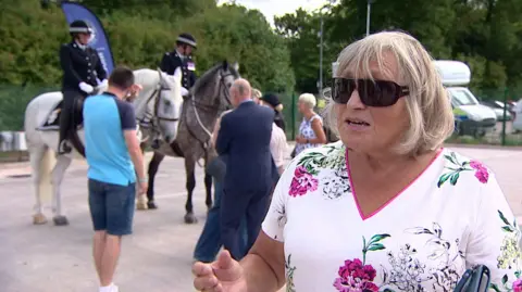 A woman wearing sunglasses stands outside speaking to a camera. A crowd of people are pictured behind her.