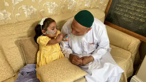 Saffanah sitting on a beige sofa with her grandfather. She is holding a toy syringe to his mouth and he is leaning down towards her.