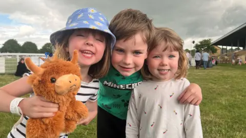 Three kids, two girls and a boy stand together smiling at the camera. The girl on the left is wearing a bucket hat and holding a brown cow soft toy. The boy, in the middle is wearing a Minecraft shirt.