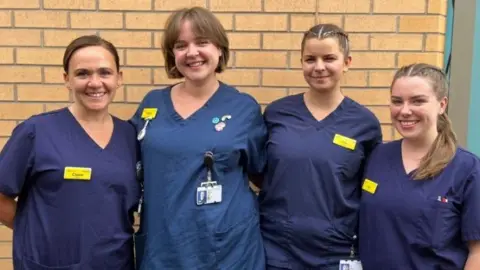 Four women who are midwives standing in front of a brick wall. They are all wearing blue scrubs and have their arms round one another's backs. They are smiling and all looking towards the camera. 