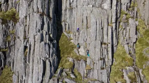Three climbers can be seen scaling the Tryfan mountain with their safety equipment - such as harnesses and hard hats - just visible around them.