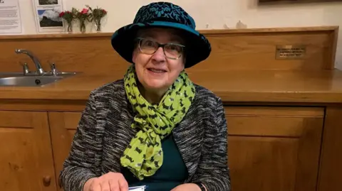 Sue Lane A smiling woman with glasses, a blue hat, green scarf and a grey cardigan. She is sat inside in front of a wooden kitchen area.