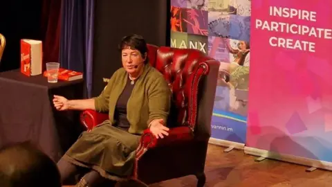 Harris sitting in a high-backed red leather armchair on a stage, with a banner behind her that says Inspire, Participate, Create.