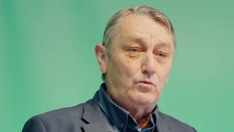 Anthony Slaughter looking to the camera, wearing a dark suit and shirt without a tie