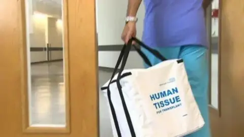 BBC A nurse wearing blue scrubs walking through a wooden doorway on a hospital corridor carrying a white bag that has the words 'human tissue for transplant' written on it in blue.
