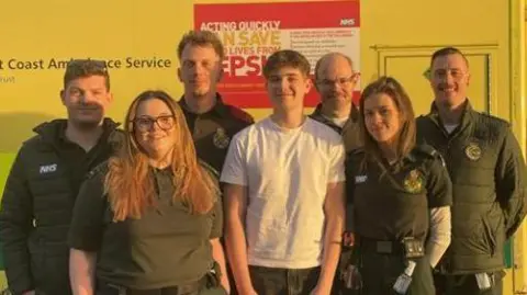 South East Coast Ambulance Service Six South East Coast Ambulance crew members standing around a teenage boy who is wearing a white shirt and grey jeans. They are standing in front of an ambulance.