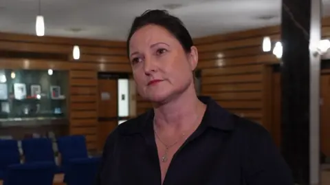 BBC A woman in a black, open-necked blouse with tied back dark hair stands in a wood-panelled room full of blue chairs.