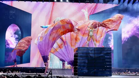 PA Media Taylor Swift performs on a platform high above Wembley stadium. Dancers are perfoming around her carrying large fabric banners.