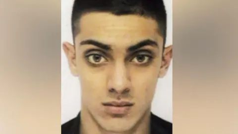 A passport-style image of a man with tidy black eyebrows, brown eyes and a narrow face. He is aged 20 and wearing a black shirt.