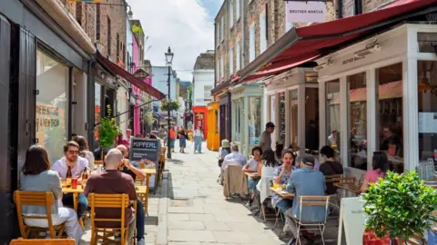 Getty Images A sunlit, eye-level shot of Camden Passage in Islington, a narrow pedestrian street lined with historic brick buildings and vibrant storefronts. Patrons dine at outdoor wooden tables under colourful awnings, while pedestrians walk through the bustling lane. Signs for local businesses hang overhead against a bright, clear sky.
