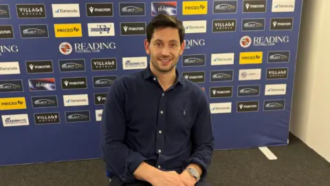 A man sat with his hands in his lap in front of a Reading Football Club sign,