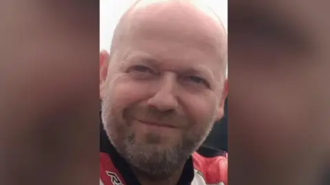 FAMILY HANDOUT A close-up of Louis, a bald man with brown stubble, smiling. He is wearing red and white racing leathers.