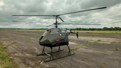 A small green unmanned helicopter on an otherwise empty airfield.