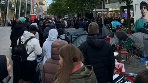 Hundreds of people wait in a queue snaking back from the shop entrance on a pedestrianised street. Glass fronted stores line the road on both sides.