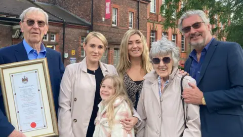 Family handout A smiling David Skentelbery and his son Gary pose with David's grey-haired wife Patricia, who wears dark glasses and wears a buff-coloured coat, and his two blond-haired granddaughters and blond-haired great-granddaughter