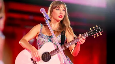 Getty Images Taylor Swift is on stage in a brilliant multicolored dress and holding a white guitar decorated with stars. She wears bright red lipstick and looks on a crowd behind a silver microphone and stands, in front of a blurred scene which is visibly lit in red.