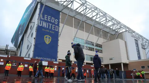 A landscape shot of Elland Road stadium's East Stand on a matchday. Banners with the club crest and name are visible. Three fans are seen at the front of the shot.