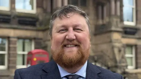 Seve Gomez Aspron has a red beard and greying hair. He is wearing a suit.
