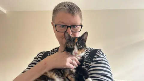 Lucy stands holding her tortoise shell cat. The cat is black, brown and white with green eyes. The cat is looking at the camera. Lucy wears a black and white striped top with black overalls. The photo is against a cream wall.