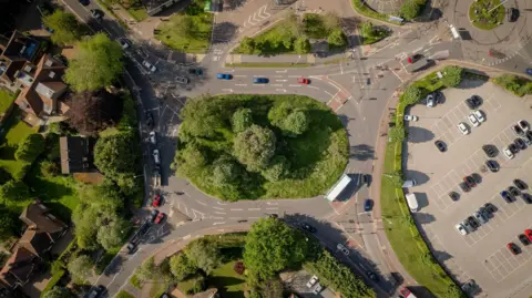 An aerial view of a roundabout with four exits and green bushes and trees in the centre.