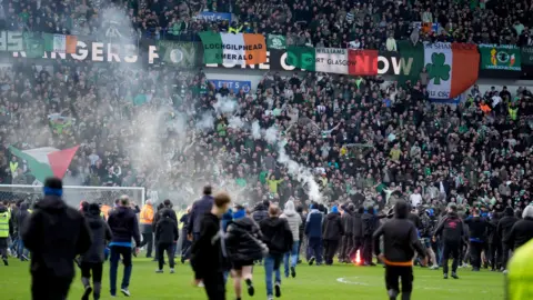 PA Media Football fans running across the pitch to confront each other. The majority are clad in black clothing. Celtic fans in the nearest stand, in Celtic tops, are visible watching on.