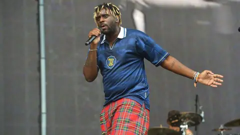 Getty Images Myles Smith, a singer with blonde dreads, wearing a blue Scotland top and red and green kilt. He is holding a microphone in one hand and has the other outstretched. He is on a stage with a drumkit behind him.