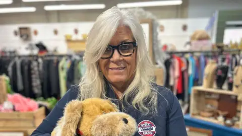 A woman called Lesley Ford smiles. She has long grey/silver hair which is down just past her shoulders. She is holding a cuddly toy dog. She is wearing a jacket with The Pet Bus logo on it. She is stood in a shop with clothes rails behind her and other items on a shelf.
