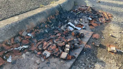 The charred remains of a box of fireworks lie on the curb of a road by the entrance of HMP Long Lartin