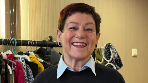 Nora Brewster has short dark hair and is wearing a black sweater with blue shirt collar, smiling at the camera. There are racks of clothes behind her. 