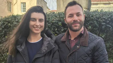 Harriet Davidson and her husband are standing in front of a green bush and smiling. They are both wearing black winter jackets. Harriet has long dark hair and her husband Adam has short brown hair and a short beard.
