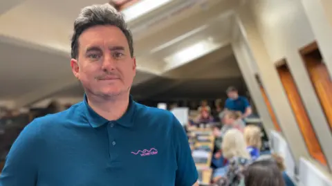 A man with dark hair is wearing a blue polo shirt with a Wavemaker logo one side. Behind him, a number of women are sat looking at laptops
