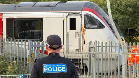A Greater Anglia train that derailed at Wickford in Essex. The train has stopped and there is damage to part of the carriage. A British Transport Police officer is standing by the train with their back to the camera.