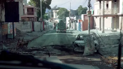 Jack Garland, BBC Burned vehicles litter a street in Port-au-Prince, an armored vehicle can be seen advancing. 