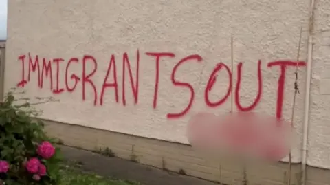 A white gable wall with the words: 'Immigrants out' sprawled in red paint. 
A section of the message has been blurred. 
A pink rose bush with some flowers can be seen at the side.  