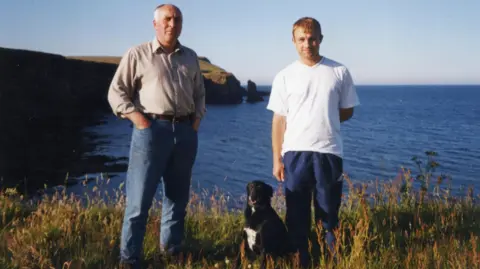 Ross family Michael Ross, his father Eddy and a black dog on top of  a cliff with the sea behind them. Ross is wearing blue trousers and a white T-shirt while Eddy is wearing blue jeans and a beige shirt. 