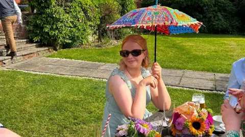 Handout Isobel, pictured sitting outside at a wooden picnic table, holding a colourful parasol above her head. She is wearing a light blue summer dress and  sunglasses. A bunch of flowers and some drinks can be seen on the table in front of her.