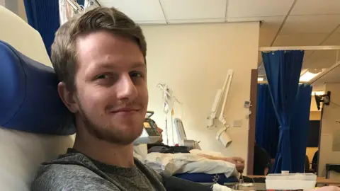 Anthony Robson has brown hair and is looking at the camera smiling. He has a short beard and moustache too. He is sitting in a hospital room with equipment and blue hospital curtains in the background. The walls are white. 