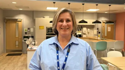 Kristie Clelland standing in front of a kitchen area, with a table to her right. She is looking directly at the camera, wearing a light blue shirt and a blue lanyard.