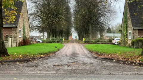 The main entrance to Whitwick Manor, along an avenue of trees in between two cottages. 