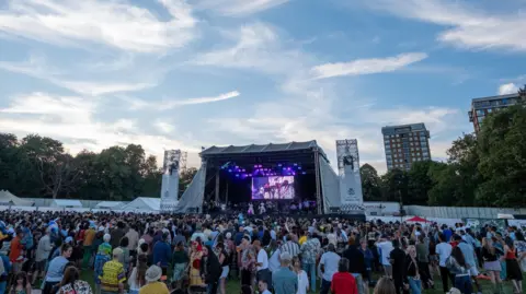 Mark McNulty Evening falls as a crowd gathers at the main stage at the Africa Oye festival. There are tall trees behind the stage, and two tower blocks loom over the scene. 