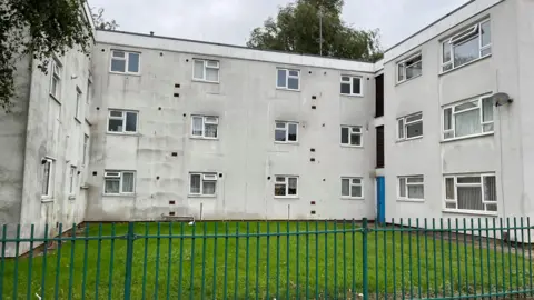 White block of flats at Alliston gardens with a green fence around the edge and grass in front