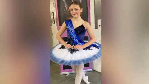 A 16-year-old girl wearing a dance uniform smiles at the camera, standing in front of a banner.